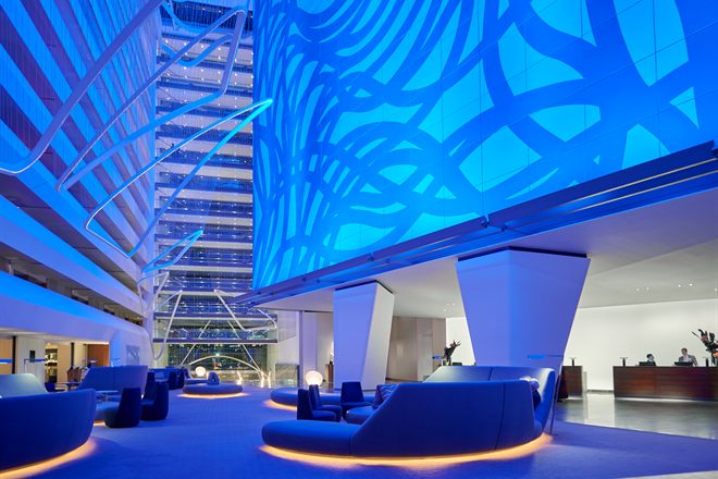 The atrium lobby at Conrad New York Downtown, lit deep blue, with a fifteen-storey looping mural rising the full height of the wall above low curved seating and the reception desk.
